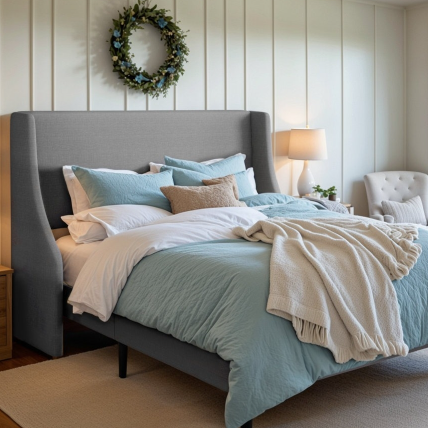 A bedroom featuring a gray upholstered bed with blue and white bedding, a wooden nightstand with lamps, an armchair, and a wall wreath. A large window offers an ocean view.
