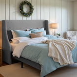 A bedroom featuring a gray upholstered bed with blue and white bedding, a wooden nightstand with lamps, an armchair, and a wall wreath. A large window offers an ocean view.