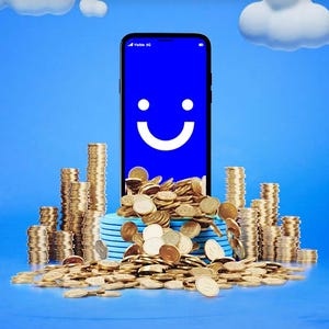 Smartphone with a smiley face on the screen amid piles of coins against a blue sky background.