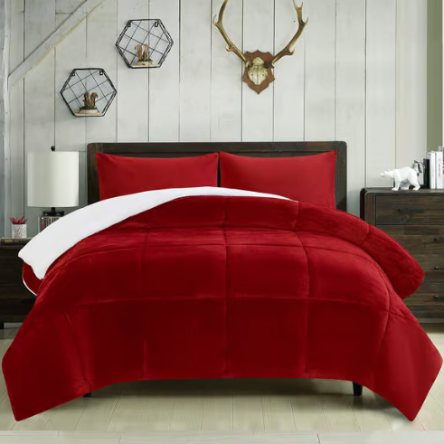 A red comforter set with reversible white lining is displayed on a bed, accompanied by matching pillows. The room decor features a wooden headboard, two modern hexagonal shelves, an antler wall mount, and a bedside table with a white lamp and decor items.