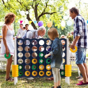 A group of people is playing with a giant Connect Four set outdoors, featuring large colored discs in a standing grid. The scene is lively with colorful decorations in the background.