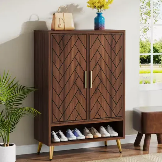 A wooden shoe cabinet with a herringbone pattern, gold handles, and legs features a top shelf for decorative items and a lower section for storing shoes. A beige handbag and a blue vase with flowers are placed on top.