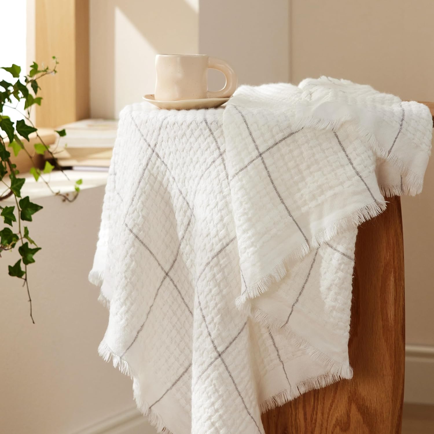 A white throw blanket with a diamond pattern and fringed edges is draped over a wooden chair back, with a cup resting on top.
