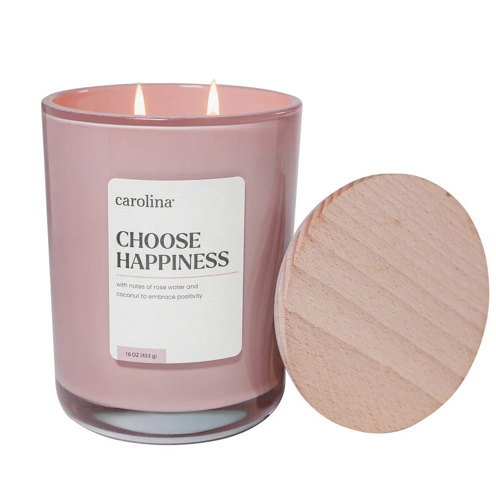 A pink glass candle labeled \“Carolina Choose Happiness\“ with notes of rose water and coconut, accompanied by a wooden lid. Capacity: 16 oz (453 g).