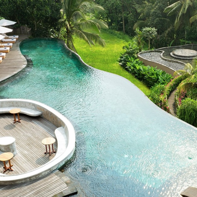 An infinity pool with wooden deck seating features umbrella-shaded loungers and lush greenery, offering a tropical, relaxing atmosphere.
