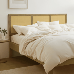 A cozy bed with cream-colored bedding, accompanied by a wooden headboard, a side table with a potted plant, and a floor lamp, all against a light-colored wall.