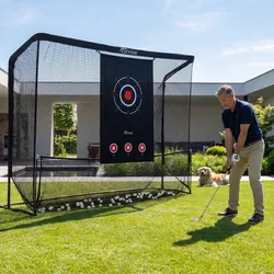 A man practices golf in his backyard using an Oxygïe golf practice net with a target. A dog lies nearby on the grass.