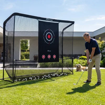 A man practices golf in his backyard using an Oxygïe golf practice net with a target. A dog lies nearby on the grass.