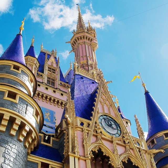 A fairy-tale castle with blue conical rooftops and ornate golden details stands majestically under a bright blue sky.