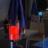 A table setup features a glowing red cube light beside bottles of liquor, a wine glass, and a candle holder; another table in the background has a glowing blue cube light.