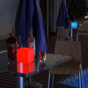 A table setup features a glowing red cube light beside bottles of liquor, a wine glass, and a candle holder; another table in the background has a glowing blue cube light.