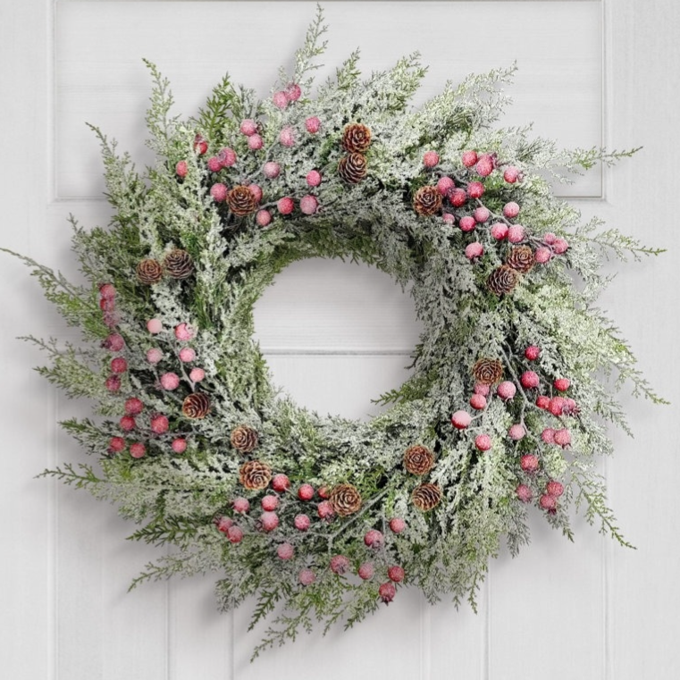A decorative wreath with snowy greenery, pink berries, and pinecones hangs on a white door next to a lantern-style wall light.