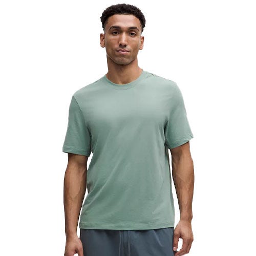 A man is wearing a light green short-sleeve shirt, featuring a simple, relaxed fit. The shirt is paired with gray shorts.