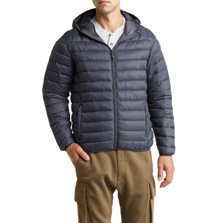 A man is wearing a navy blue quilted jacket with a hood, a zip front, and two side pockets, paired with khaki trousers.