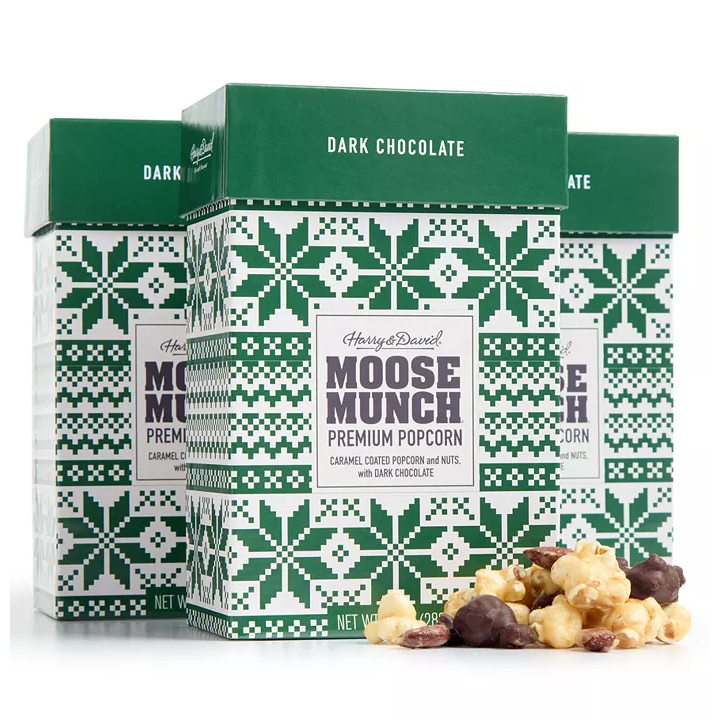 Three boxes of Harry & David's Moose Munch Premium Popcorn in dark chocolate flavor, featuring caramel-coated popcorn and nuts, decorated with green holiday-themed patterns. Some popcorn pieces are visible in front of the boxes.