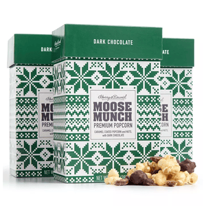 Three boxes of Harry & David's Moose Munch Premium Popcorn in dark chocolate flavor, featuring caramel-coated popcorn and nuts, decorated with green holiday-themed patterns. Some popcorn pieces are visible in front of the boxes.