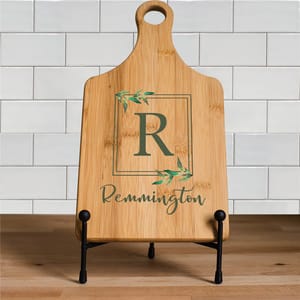A bamboo paddle cutting board featuring a large letter \“R\“ and the name \“Remmington,\“ adorned with green leaf accents, stands on a wooden countertop against a white tiled backsplash.