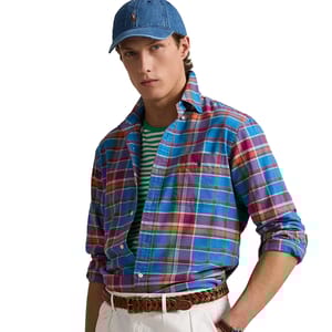 A person is wearing a blue plaid button-up shirt over a green and white striped tee, paired with a woven brown belt and white pants, accessorized with a blue denim cap.