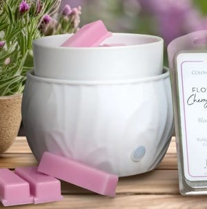 A white wax warmer with pink wax melts sits alongside a pack labeled \“Cherry Blossom,\“ against a backdrop of flowers.