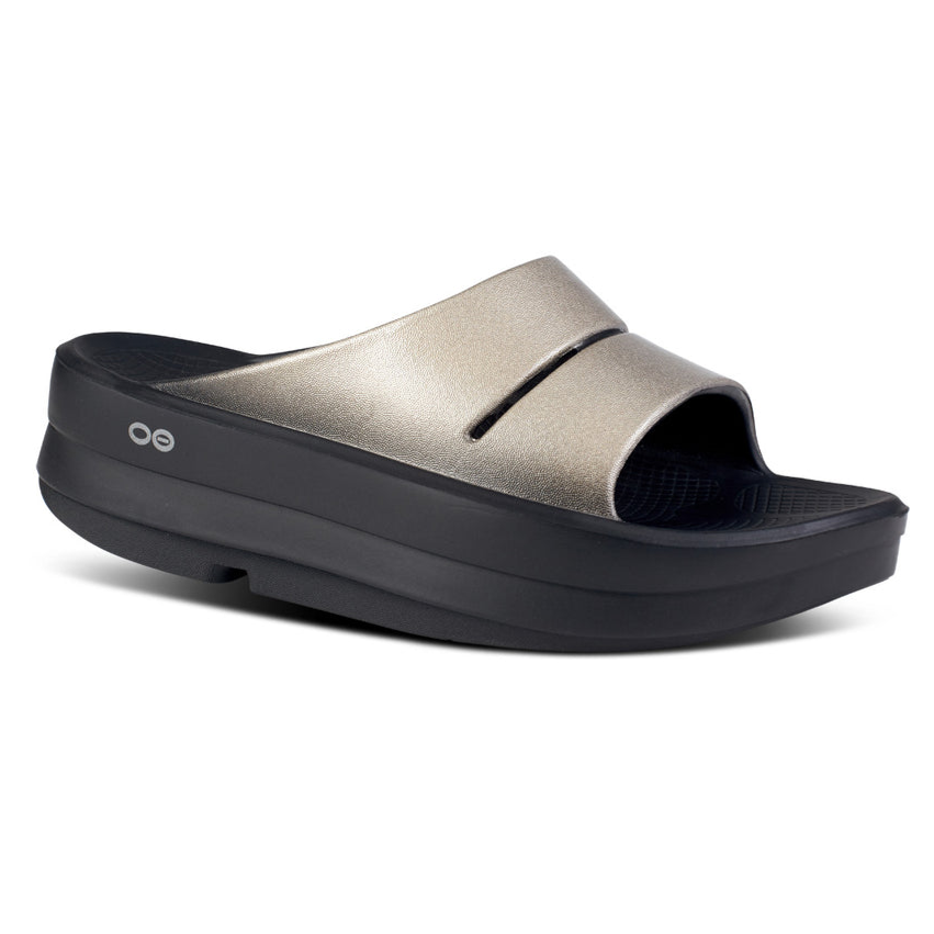 A metallic gold open-toe slide sandal with a thick black sole, featuring a simple design and small circular logo on the side.