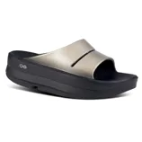 A metallic gold open-toe slide sandal with a thick black sole, featuring a simple design and small circular logo on the side.