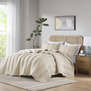 A beige comforter set with coordinating shams on a bed, accompanied by a bedside table, lamp, rug, and wall art.