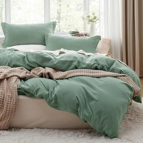 A bed with green bedding, including a comforter and pillows, is accompanied by a textured beige throw blanket. The setting features a window with sheer curtains allowing natural light to filter in.