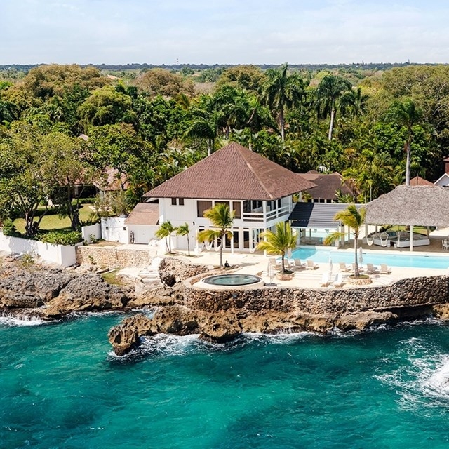 A beachfront villa with a brown roof and large windows is surrounded by lush greenery. It features an infinity pool and hot tub overlooking the ocean, set on rocky terrain with palm trees onsite.