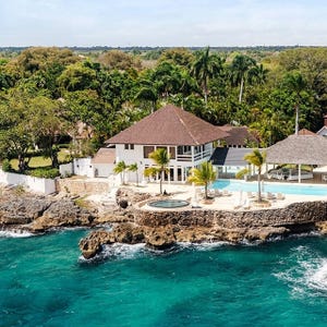 A beachfront villa with a brown roof and large windows is surrounded by lush greenery. It features an infinity pool and hot tub overlooking the ocean, set on rocky terrain with palm trees onsite.