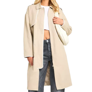 A beige trench coat with a relaxed fit, featuring white piping accents and long sleeves. It is paired with a white crop top and gray jeans, complemented by a white woven shoulder bag.