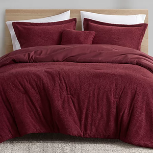 A bed features a burgundy bedding set, which includes a duvet cover, two pillow shams, and a decorative cushion on a woven rug.
