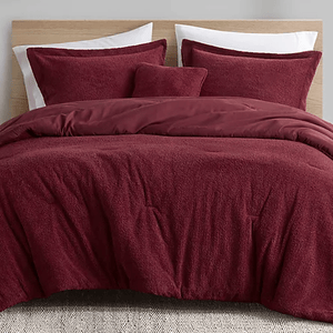 A bed features a burgundy bedding set, which includes a duvet cover, two pillow shams, and a decorative cushion on a woven rug.