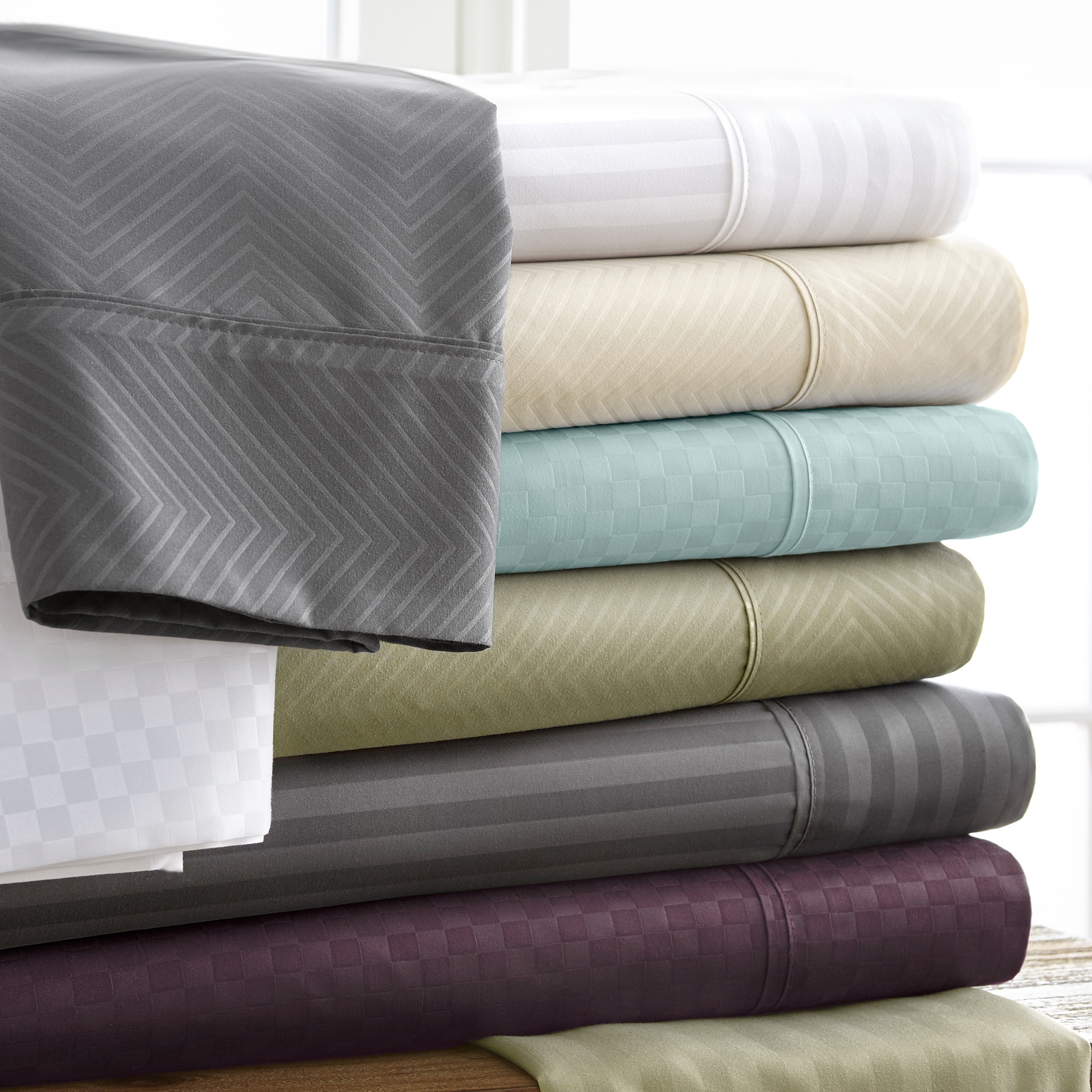 Stack of folded bed sheets in various colors, including gray, white, beige, turquoise, green, and purple, featuring different textured patterns such as chevron and checkered designs.