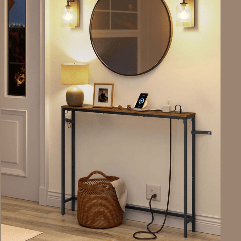 A narrow console table with a rustic wood top and metal legs is displayed beneath a round mirror. It holds a lamp, a framed photo, and a smartphone dock. Two wall sconces and a woven basket are nearby on a wooden floor.