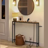 A narrow console table with a rustic wood top and metal legs is displayed beneath a round mirror. It holds a lamp, a framed photo, and a smartphone dock. Two wall sconces and a woven basket are nearby on a wooden floor.