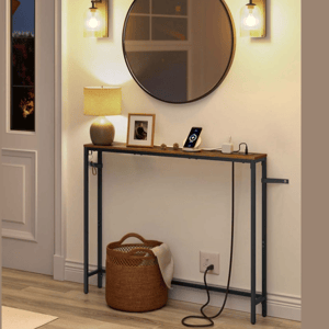 A narrow console table with a rustic wood top and metal legs is displayed beneath a round mirror. It holds a lamp, a framed photo, and a smartphone dock. Two wall sconces and a woven basket are nearby on a wooden floor.