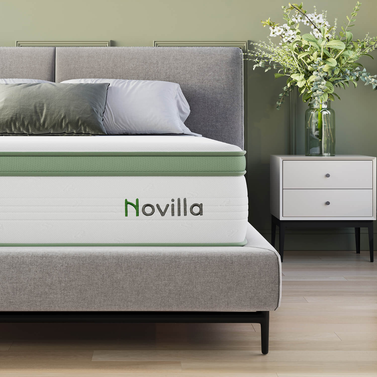 A gray bed with a green-accented Novilla mattress is shown alongside a white nightstand with a vase of green and white flowers.