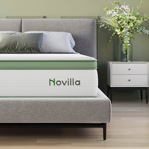 A gray bed with a green-accented Novilla mattress is shown alongside a white nightstand with a vase of green and white flowers.