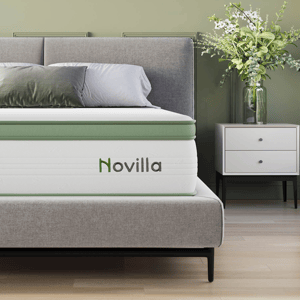 A gray bed with a green-accented Novilla mattress is shown alongside a white nightstand with a vase of green and white flowers.