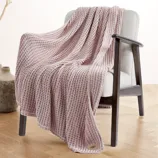 A light pink knit throw blanket draped over a light gray upholstered chair with wooden armrests, featuring a small label at the corner with \“UGG\“ on it, on a wooden floor.