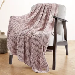 A light pink knit throw blanket draped over a light gray upholstered chair with wooden armrests, featuring a small label at the corner with \“UGG\“ on it, on a wooden floor.