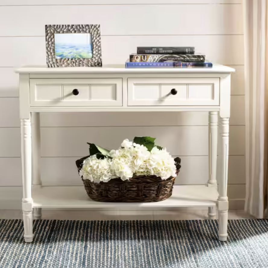 A white console table with two drawers features a wicker basket of white flowers on the lower shelf, and a collection of books and a photo frame on top.