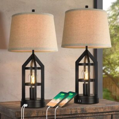 Two table lamps with geometric black frames and beige shades are on a wooden surface, each featuring built-in USB charging ports with two smartphones connected.