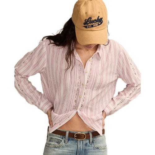 A person is wearing a light pink striped button-up shirt, blue jeans, and a tan cap with \“Lucky EST. 1990\“ written on it.