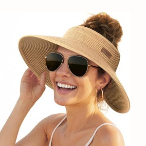 Wide-brimmed sun visor hat in a light tan color, with a strap for securing, worn by a woman with sunglasses and hoop earrings.