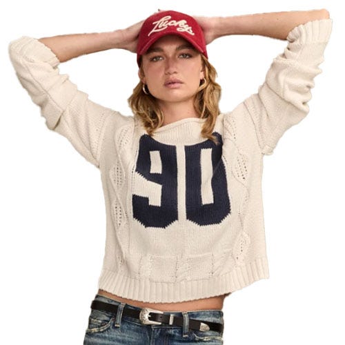A person wears a cream sweater with a large \“90\“ design, blue jeans, a black belt, and a red baseball cap.