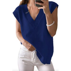 A woman wearing a blue sleeveless ribbed V-neck sweater paired with white pants and a bracelet. She is holding a phone in one hand.
