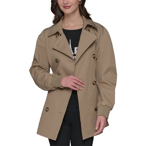 A woman wears a tan double-breasted trench coat with large buttons and ribbed cuffs over a black top and black pants.