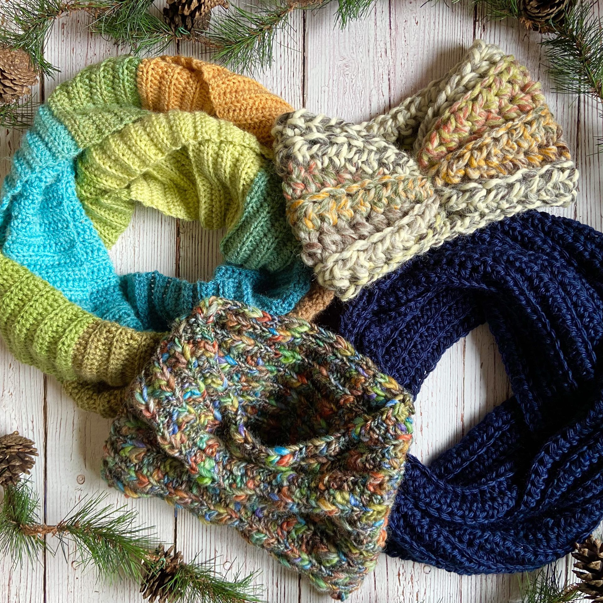 Four knitted scarves in various colors and patterns are arranged on a white wooden background adorned with pine branches and cones.