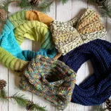 Four knitted scarves in various colors and patterns are arranged on a white wooden background adorned with pine branches and cones.